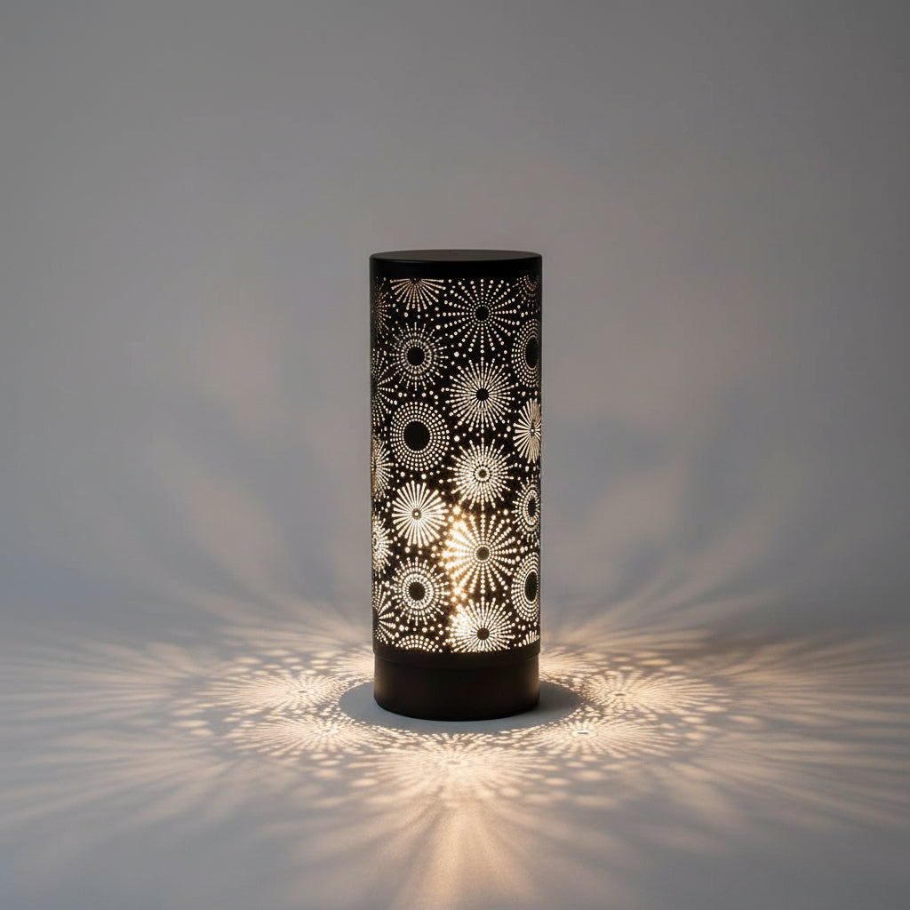 A black cylindrical table lamp with a glowing white laser-cut geometric starburst pattern, set on a wooden surface against a geometric tiled wall with a small potted succulent on the left.