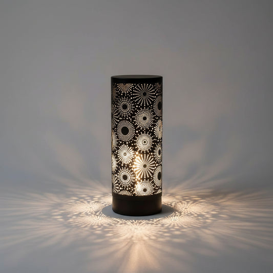 A black cylindrical table lamp with a glowing white laser-cut geometric starburst pattern, set on a wooden surface against a geometric tiled wall with a small potted succulent on the left.