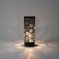 A black cylindrical table lamp with a glowing white laser-cut geometric starburst pattern, set on a wooden surface against a geometric tiled wall with a small potted succulent on the left.